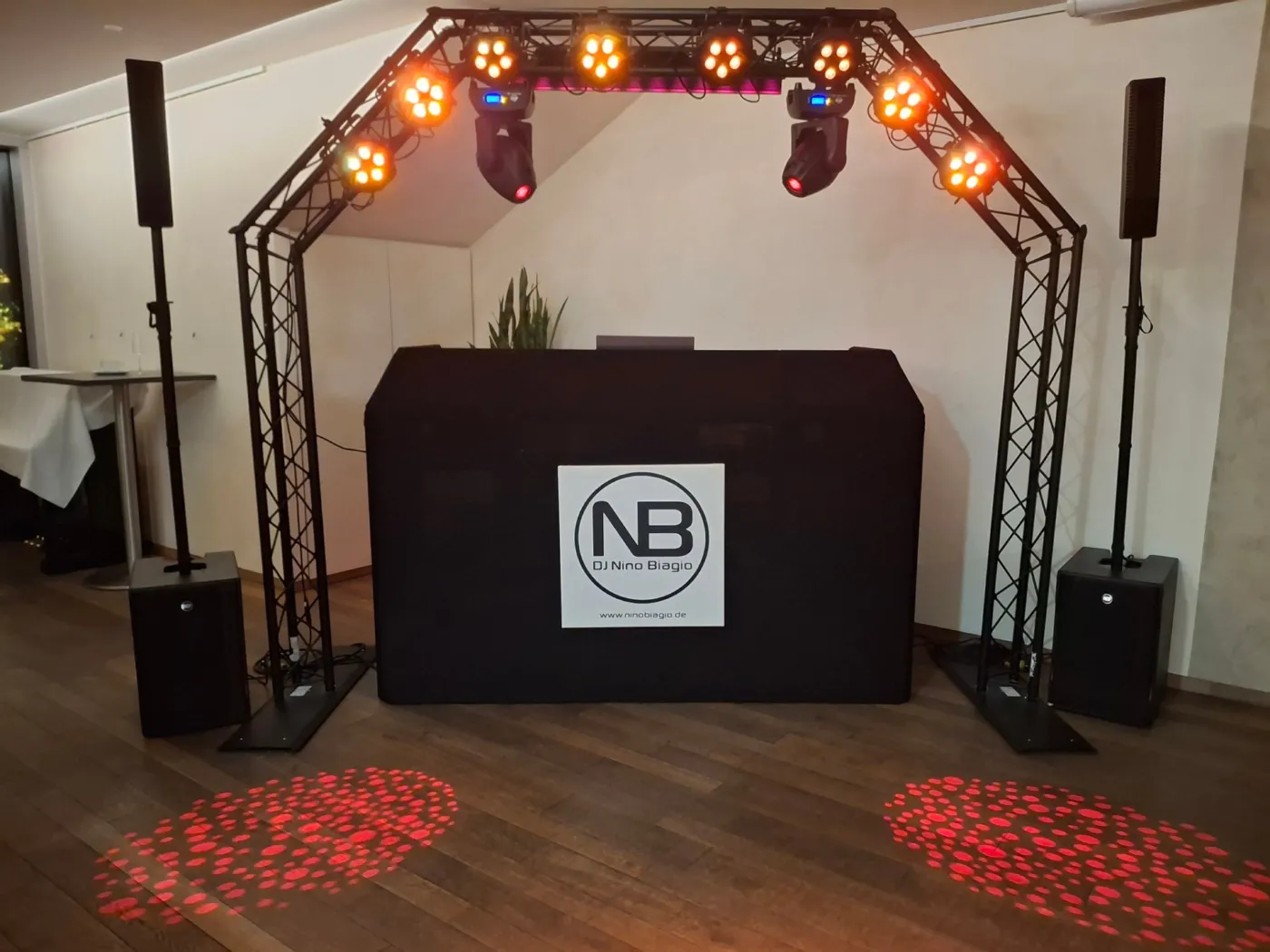 DJ-Setup im Eventhaus Waldbronn: Traversenbogen mit orangem Licht, rote Lichtmuster auf dem Holzboden, schwarzes DJ-Pult mit Logo zwischen zwei Lautsprechern