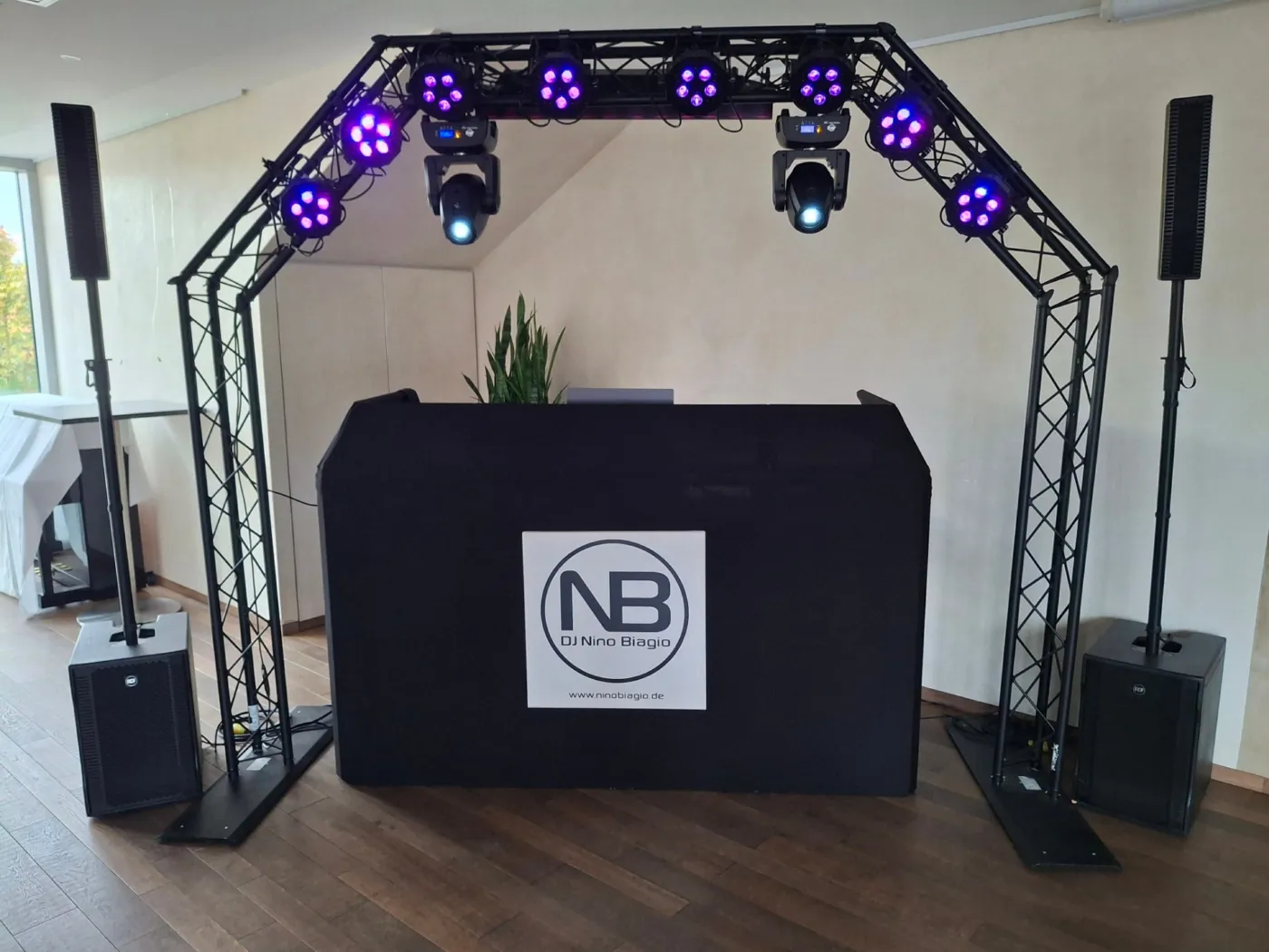 DJ-Setup im Eventhaus Waldbronn bei Tageslicht: Traversenbogen mit violettem Licht und Movingheads, schwarzes DJ-Pult mit Logo