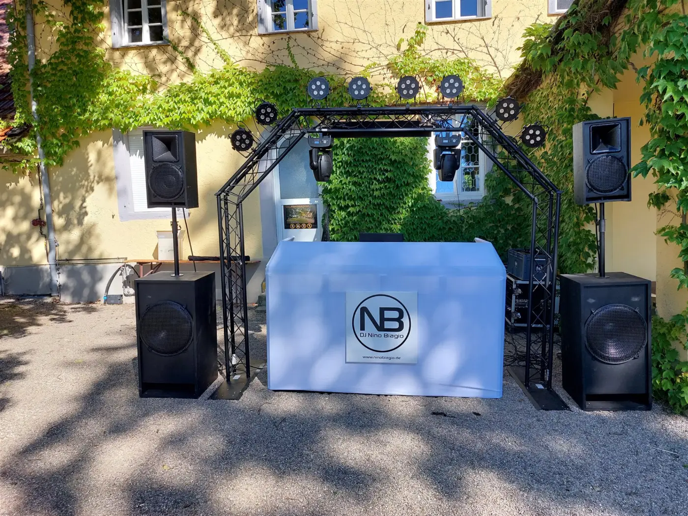 DJ-Setup von Nino Biagio im Hof einer Hochzeitslocation: Traversenbogen mit Movingheads, Subwoofer und weißes DJ-Pult mit Logo